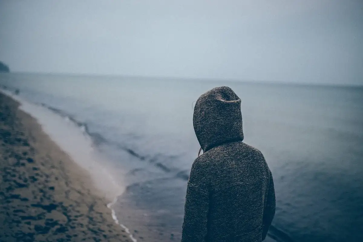 A person seen from behind in a gray knit hoodie, hood up, facing a calm lake shore
