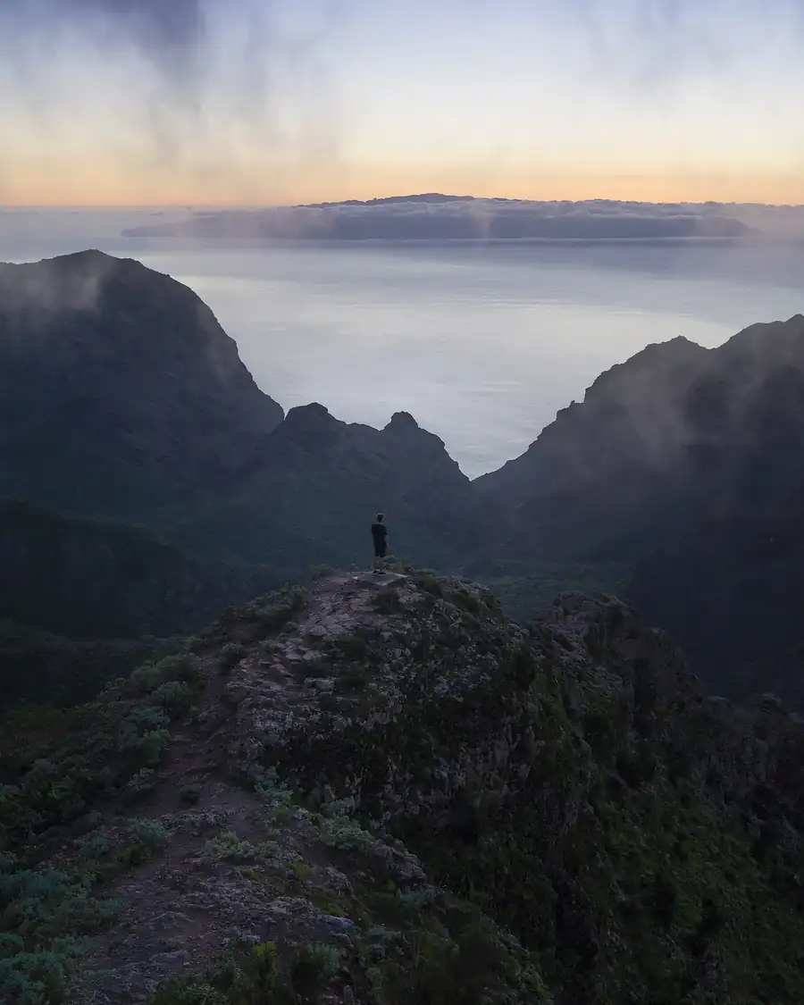 A hiker small on a rocky lookout above folded mountains and a sea of cloud at first light