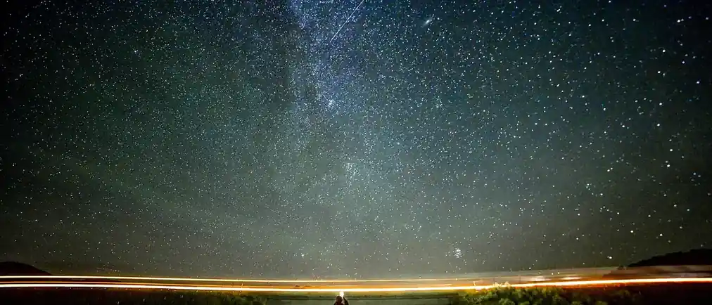 A night sky packed with stars above a dark valley, a thin line of light along the horizon