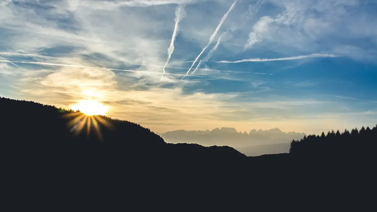 The sun setting behind a dark forested ridge under a sky streaked with thin cloud